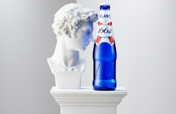 Kronenbourg Blanc bottle displayed beside a classical statue in a staged gallery setting.